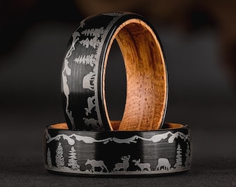 May include: Two black tungsten carbide rings with a wood inlay. The rings are engraved with a nature scene featuring trees, mountains, and animals.