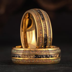 Pode incluir: Duas alianças de casamento douradas com uma incrustação central de flocos pretos e dourados e uma faixa circundante de material iridescente semelhante ao opala. Os anéis são exibidos em um fundo escuro, destacando seu acabamento dourado polido e design intrincado.