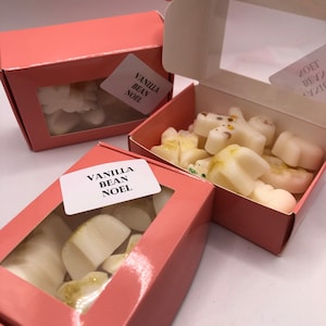 Könnte beinhalten: Drei rosa Schachteln mit transparenten Fenstern, die jeweils weiße Wachsschmelzen enthalten. Die Schachteln tragen Etiketten mit der Aufschrift "VANILLA BEAN NOEL". Die Wachsschmelzen haben eine leichte Bestäubung mit goldenem Glitzer.