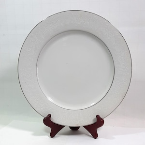 Puede incluir: Un plato de cerámica blanco con un borde plateado y un patrón floral en relieve alrededor del borde. El plato se exhibe sobre un soporte de madera rojo oscuro. El plato es redondo y tiene una superficie lisa y brillante.