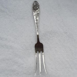 May include: A silver-coloured metal souvenir fork featuring a three-pronged head and a decorative handle. The handle displays an embossed image of a sailing ship at the top, followed by the text 1620 and the word PLYMOUTH written vertically.
