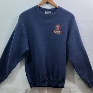 May include: A navy blue sweatshirt with a crew neck and long sleeves. The sweatshirt features a small graphic on the chest with the text "Cheers" in a stylized font. The graphic includes a red and brown design.