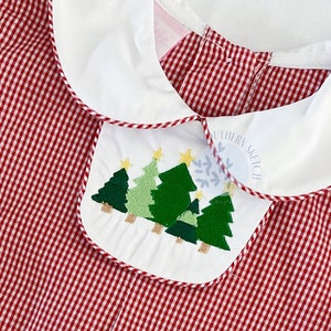 May include: Red and white gingham dress with a white Peter Pan collar and a green embroidered Christmas tree design on the front. The dress has short sleeves and a tag that says "Petit Bebe".