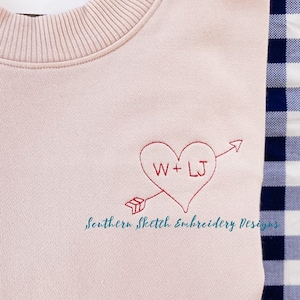 May include: A pale pink sweatshirt with a red embroidered heart that says "W + LJ" and an arrow pointing to the right. The text "Southern Sketch Embroidery Designs" is embroidered below the heart.