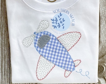 Op de afbeelding: Twee witte T-shirts met een blauwe en witte vliegtuigapplicatie. Het vliegtuig heeft een ruitjespatroon en een grijze neus. De shirts hebben ook een blauwe cirkel met de tekst "Southern Sketch" en een klein groen bladerenontwerp.