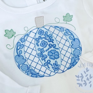 Puede incluir: Body blanco de manga larga con un diseño de calabaza bordada en azul. La calabaza presenta un patrón de celosía y detalles florales. Hojas verdes y un tallo gris completan el diseño. El body tiene un puño con volantes y cierre de broche.