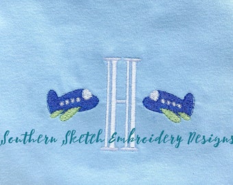 Op de afbeelding: Een lichtblauw T-shirt met een witte geborduurde letter "H" en twee blauwe vliegtuigen aan weerszijden van de letter. De tekst "Southern Sketch Embroidery Designs" is onder de letter geborduurd.