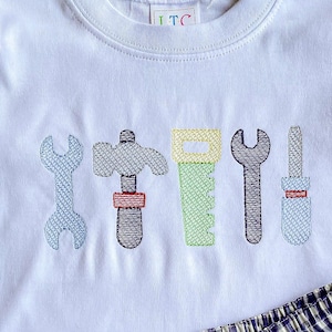 May include: White short-sleeved shirt with a row of embroidered tools, including a wrench, hammer, saw, screwdriver, and another wrench. The shirt is paired with blue and white gingham shorts.