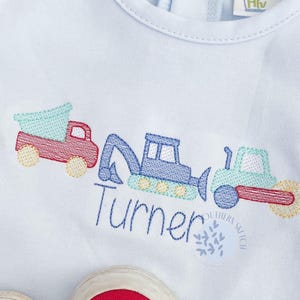 May include: A light blue sleeveless shirt with an embroidered design of construction vehicles and the name "Turner". The shirt is paired with red and white checkered shorts.