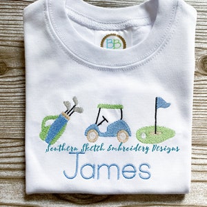 May include: White t-shirt with embroidered golf-themed design, including a golf bag, golf cart, and flag. The text "Southern Sketch Embroidery Designs" and the name "James" are also embroidered on the shirt.