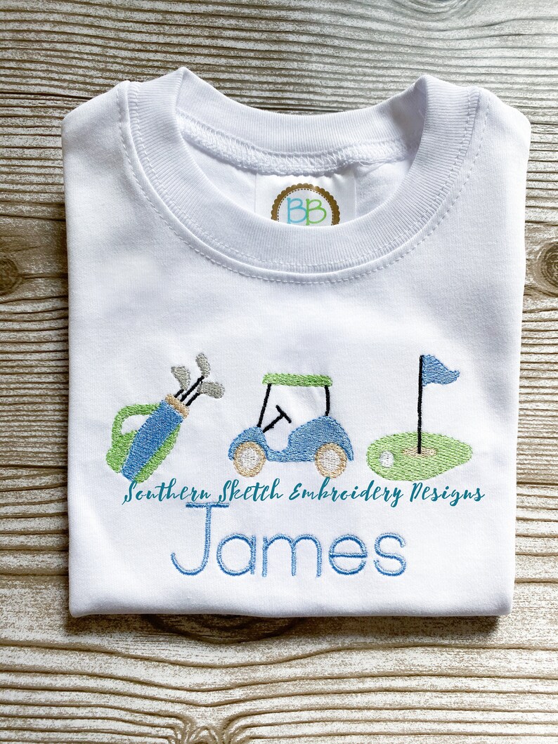 May include: White t-shirt with embroidered golf-themed design, including a golf bag, golf cart, and flag. The text "Southern Sketch Embroidery Designs" and the name "James" are also embroidered on the shirt.