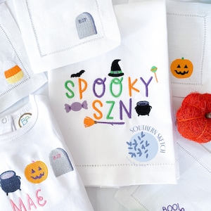 May include: White embroidered kitchen towels with Halloween themes. One towel features a witch's hat, candy corn, a cauldron, a broom, and the text "Spooky Szn". Another towel features a pumpkin, a tombstone, and the text "Boo Boo Boo".