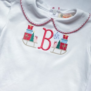 Puede incluir: Camiseta blanca de manga larga con cuello Peter Pan rojo y blanco. La camiseta tiene una letra "B" bordada en rojo con una escena navideña de regalos y un árbol en un trineo.