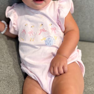 May include: A light pink baby bodysuit with ruffled sleeves. The bodysuit features an embroidered design of four geese wearing pink bows. The text "SOUTHERN SKETCH" is printed on the front of the bodysuit.
