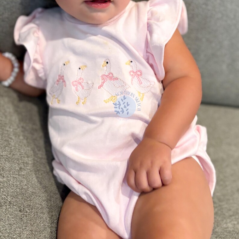 May include: A light pink baby bodysuit with ruffled sleeves. The bodysuit features an embroidered design of four geese wearing pink bows. The text "SOUTHERN SKETCH" is printed on the front of the bodysuit.