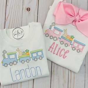 May include: Two white shirts with embroidered Easter train designs. The shirt on the left has the name Landon embroidered below the train. The shirt on the right has the name Alice embroidered below the train.
