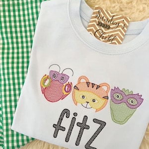 Könnte beinhalten: Ein hellblaues T-Shirt mit gestickten Bildern eines Krebses, eines Tigers und eines Monsters mit Maske. Der Text "Fitz" ist unter den Bildern gestickt. Das T-Shirt liegt auf einem grün-weiß karierten Stoff.