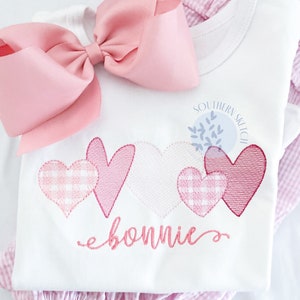 以下が含まれることがあります： ピンクのギンガムチェックのハートと「bonnie」という名前が刺繍された白いロンパース。ロンパースはピンクのギンガムチェックのスカートと大きなピンクのリボンと組み合わせています。