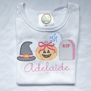 Pode incluir: Camiseta branca para criança com design bordado de Halloween em rosa e cinza. O design inclui um chapéu de bruxa, uma abóbora com um laço e uma lápida com a palavra "RIP". O nome "Adelaide" está bordado abaixo do design.