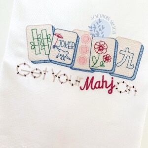 May include: White embroidered towel featuring colorful Mahjong tiles with bamboo, a joker, flowers, and Chinese characters. The text "Get Your Mahj On" is embroidered below the tiles in a playful font.