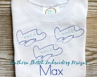 Op de afbeelding: Een witte baby romper met blauwe geborduurde vliegtuigen en de naam "Max" eronder. De romper heeft een klein label met de letters "BB" erop.