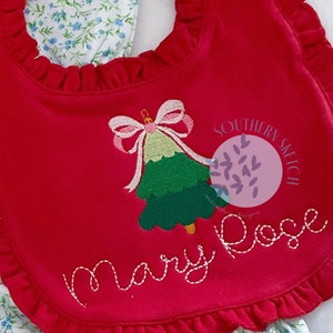 May include: Red Christmas-themed bib with a green embroidered Christmas tree, a pink bow, and the text "Mary Rose" embroidered below. The bib has a ruffled edge and the text "Southern Sketch" is embroidered in a circle on the right side.