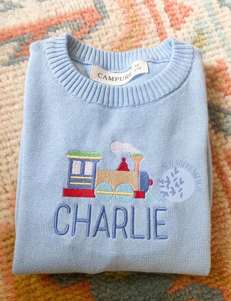 May include: A light blue knitted sweater with a colorful embroidered train and the name "CHARLIE" on the front. The sweater has a Campure tag.