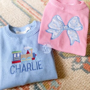 May include: Two children's sweaters, one light blue with an embroidered train and the name "CHARLIE", the other pink with a blue and white bow.