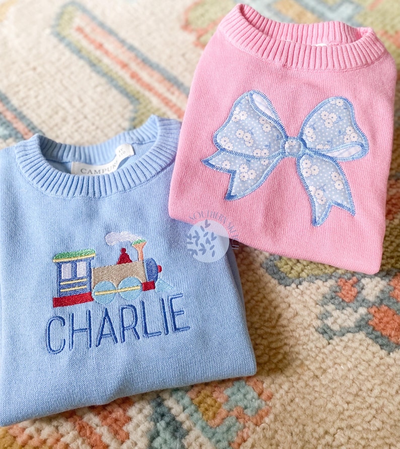 May include: Two children's sweaters, one light blue with an embroidered train and the name "CHARLIE", the other pink with a blue and white bow.