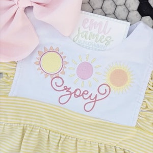 May include: A white dress with yellow and white stripes and a white bodice. The bodice has three embroidered suns in pink, yellow, and orange. The name "Zoey" is embroidered in pink on the bodice. The dress is for a baby girl.