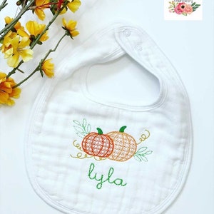 May include: White baby bib with embroidered pumpkins and green leaves. The name "Lyla" is embroidered below the pumpkins.