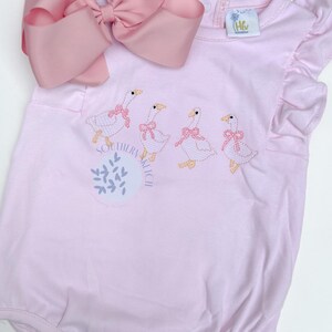 May include: Pink baby romper with embroidered geese wearing pink bows. The romper has a large pink bow at the top and the words "Southern Kitsch" are printed on the front. The romper has ruffle sleeves.