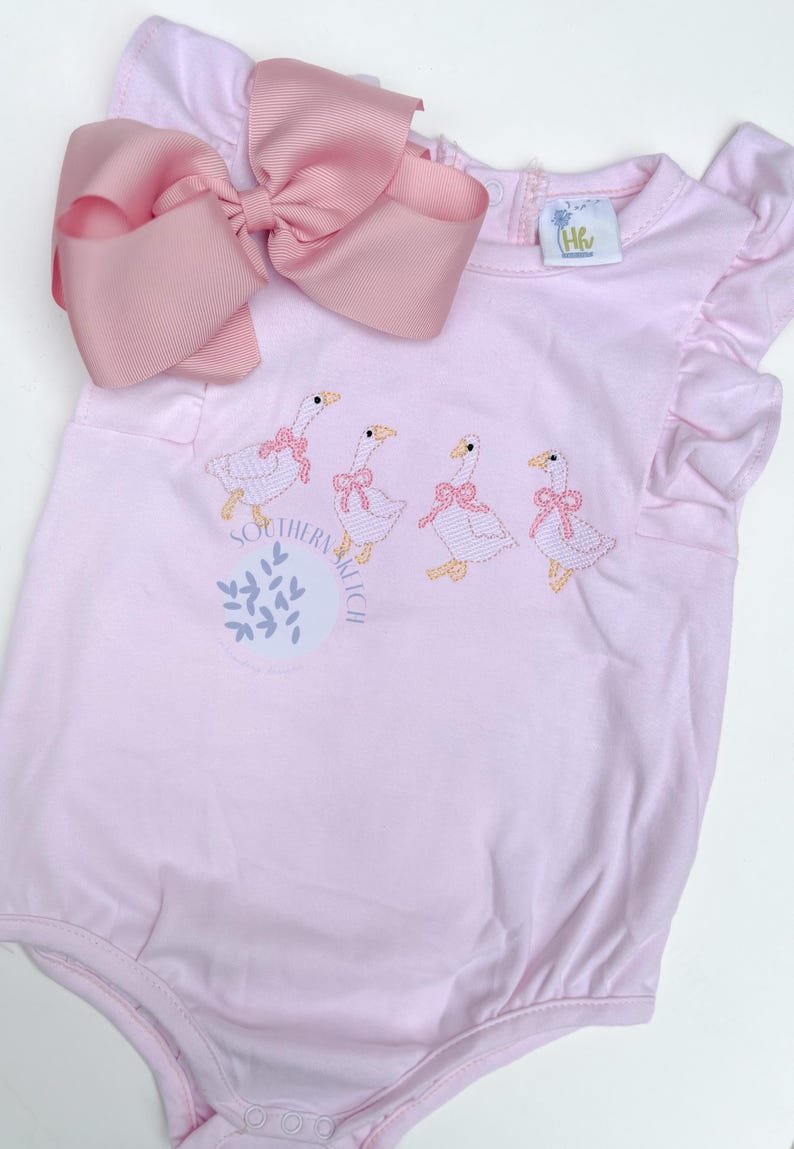 May include: Pink baby romper with embroidered geese wearing pink bows. The romper has a large pink bow at the top and the words "Southern Kitsch" are printed on the front. The romper has ruffle sleeves.