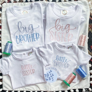 May include: Four white t-shirts with embroidered text. Two shirts say "big brother" and "big sister" in pink and blue script. Two shirts say "little sister" and "little brother" in pink and blue script.