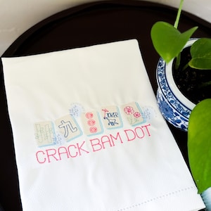 Puede incluir: Toalla blanca bordada con el texto "CRACK BAM DOT" en rosa. La toalla presenta fichas de Mahjong bordadas en azul claro, rosa y verde. Una planta decorativa en una maceta azul y blanca est&aacute; en el fondo.