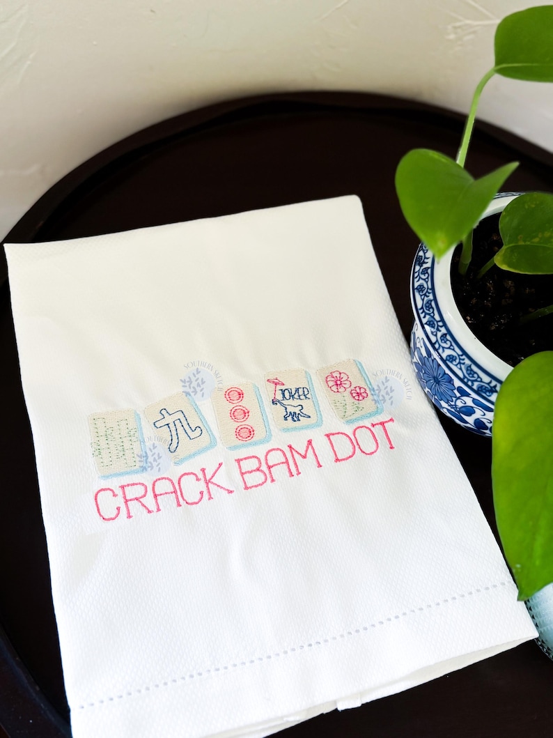 Puede incluir: Toalla blanca bordada con el texto "CRACK BAM DOT" en rosa. La toalla presenta fichas de Mahjong bordadas en azul claro, rosa y verde. Una planta decorativa en una maceta azul y blanca est&aacute; en el fondo.