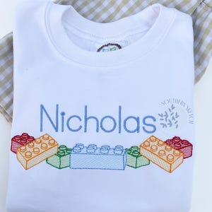May include: White t-shirt with a blue embroidered name "Nicholas" and a row of colorful embroidered toy building blocks. The shirt also has a small embroidered logo that says "Southern Stitch".