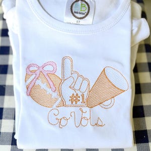 May include: White t-shirt with embroidered orange and pink football, #1 foam finger, and megaphone. The text "Go Vols" is embroidered in orange script. A tag on the shirt reads "Blanks Boutique".