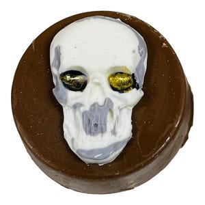 Puede incluir: Un dulce cubierto de chocolate con un dise&ntilde;o de calavera blanca. La calavera tiene detalles grises y detalles dorados en los ojos. El dulce es redondo y tiene una base marr&oacute;n oscuro.