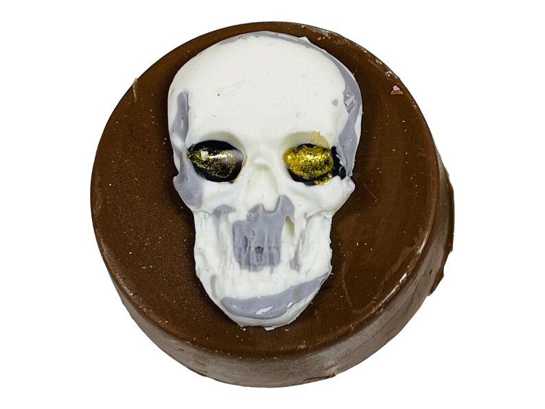 Puede incluir: Un dulce cubierto de chocolate con un dise&ntilde;o de calavera blanca. La calavera tiene detalles grises y detalles dorados en los ojos. El dulce es redondo y tiene una base marr&oacute;n oscuro.