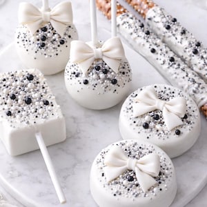 Puede incluir: Una variedad de dulces cubiertos de chocolate blanco, que incluyen cake pops, galletas y palitos de pretzel. Los postres est&aacute;n decorados con chispas negras, plateadas y blancas, y adornados con lazos blancos. Dispuestos sobre una superficie de m&aacute;rmol blanco.