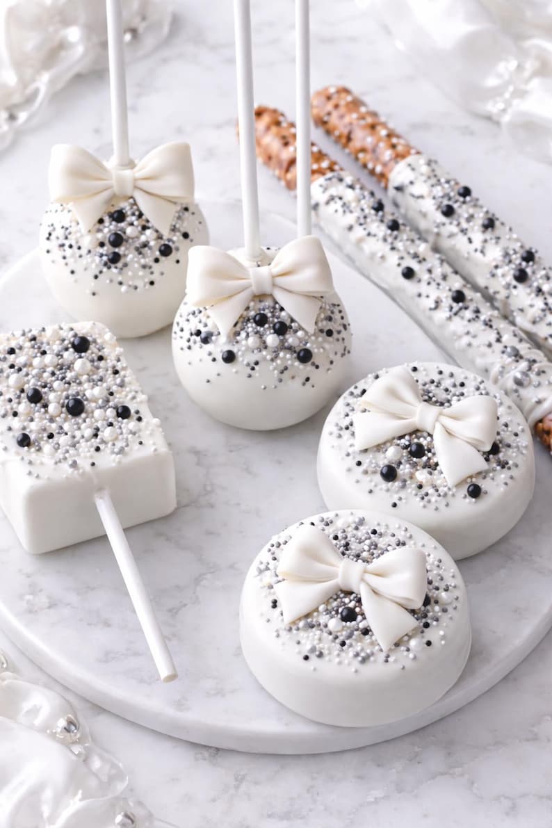 Puede incluir: Una variedad de dulces cubiertos de chocolate blanco, que incluyen cake pops, galletas y palitos de pretzel. Los postres est&aacute;n decorados con chispas negras, plateadas y blancas, y adornados con lazos blancos. Dispuestos sobre una superficie de m&aacute;rmol blanco.