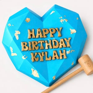 May include: A bright blue, heart-shaped treat with gold lettering that reads "HAPPY BIRTHDAY KYLAH." Gold leaf accents the surface. A wooden mallet with "BREAK ME" printed on it rests beside the treat.