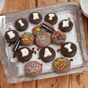 Puede incluir: Galletas s&aacute;ndwich cubiertas de chocolate dispuestas en una bandeja transparente. Algunas galletas tienen letras blancas que deletrean "BIRTHAY", mientras que otras est&aacute;n decoradas con chispas y caramelos de colores. Las galletas est&aacute;n sobre una superficie de madera.