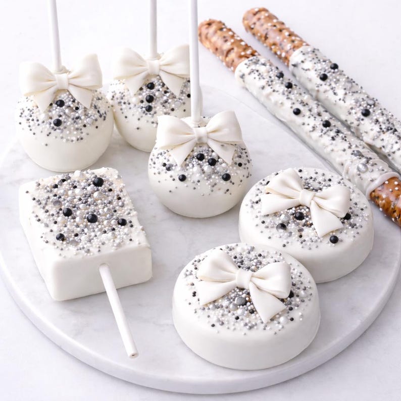 Puede incluir: Una variedad de dulces cubiertos de chocolate blanco, que incluyen cake pops, galletas y pretzels, decorados con chispas plateadas y negras y lazos blancos. Los dulces est&aacute;n dispuestos en una fuente de m&aacute;rmol blanco.