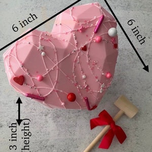 May include: A pink chocolate heart-shaped treat decorated with white and pink sprinkles, red hearts, and pink and white candy. The heart is 6 inches wide and 3 inches tall. A wooden mallet with a red bow is included.