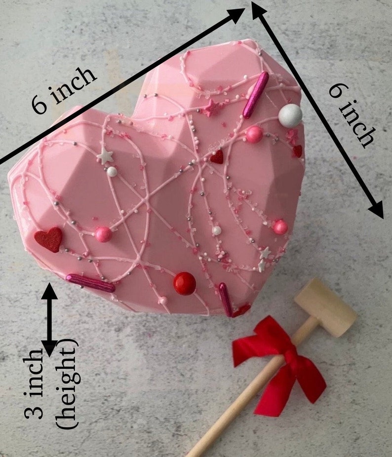 May include: A pink chocolate heart-shaped treat decorated with white and pink sprinkles, red hearts, and pink and white candy. The heart is 6 inches wide and 3 inches tall. A wooden mallet with a red bow is included.