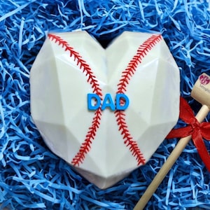 K&ouml;nnte beinhalten: Ein wei&szlig;er Schokoladen-Baseball-Herz mit roten N&auml;hten und dem Wort "DAD" in blauen Buchstaben. Ein Holzhammer mit einem roten Band ist im Hintergrund.