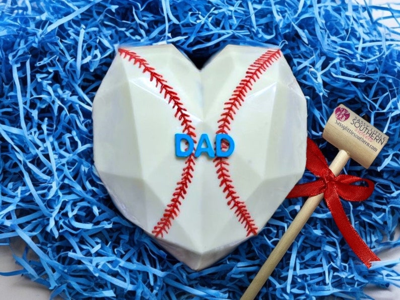 K&ouml;nnte beinhalten: Ein wei&szlig;er Schokoladen-Baseball-Herz mit roten N&auml;hten und dem Wort "DAD" in blauen Buchstaben. Ein Holzhammer mit einem roten Band ist im Hintergrund.