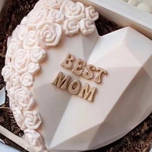 May include: A white heart-shaped cake decorated with white roses and the words "BEST MOM" in gold letters.
